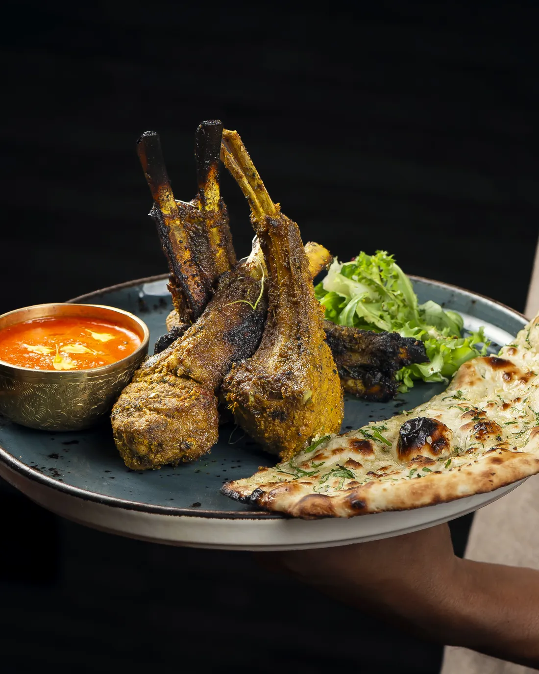 A plate of lamb chops, naan and sauce, held in one hand against a dark background