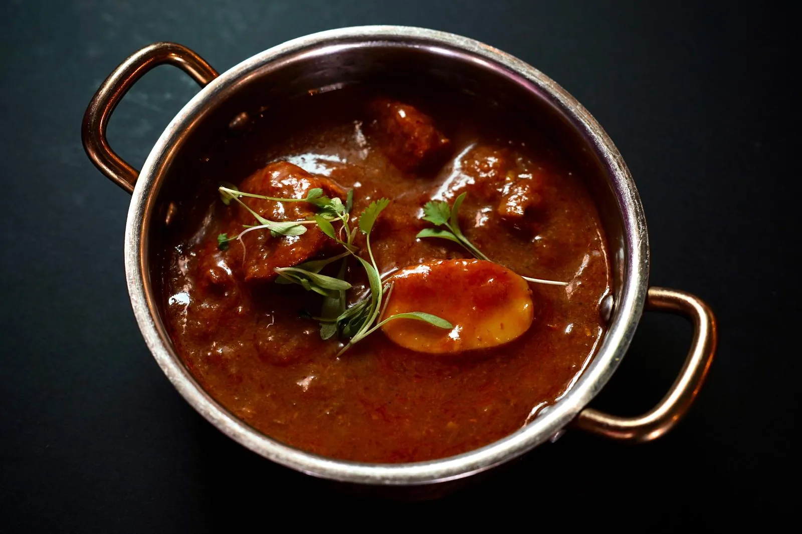 Railway lamb curry in a copper pan seen from above: pieces of lamb and a potato in a dark sauce, with coriander sprouts
