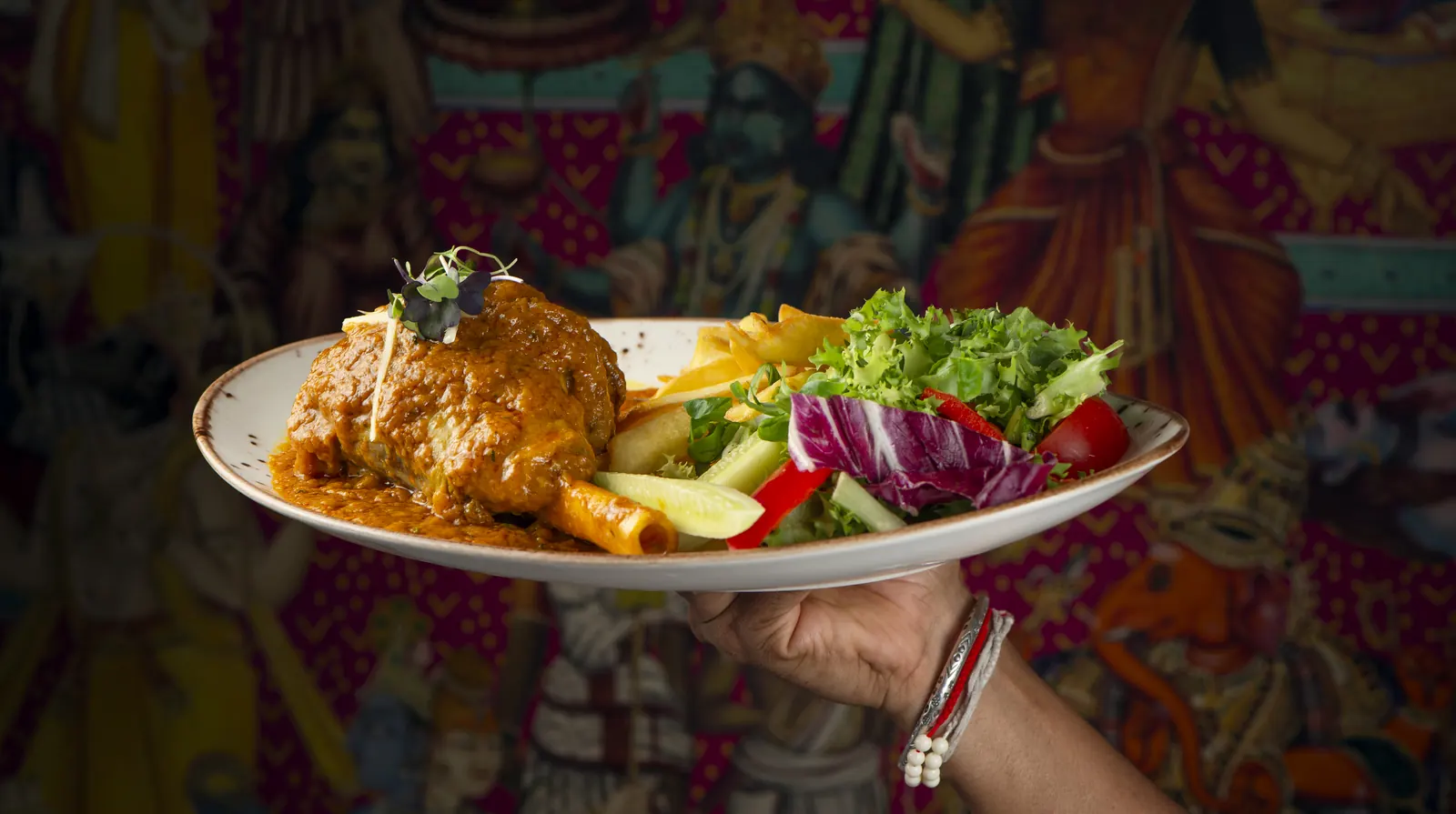 A plate of lamb in sauce with salad, held up in front of a colourful Indian mural
