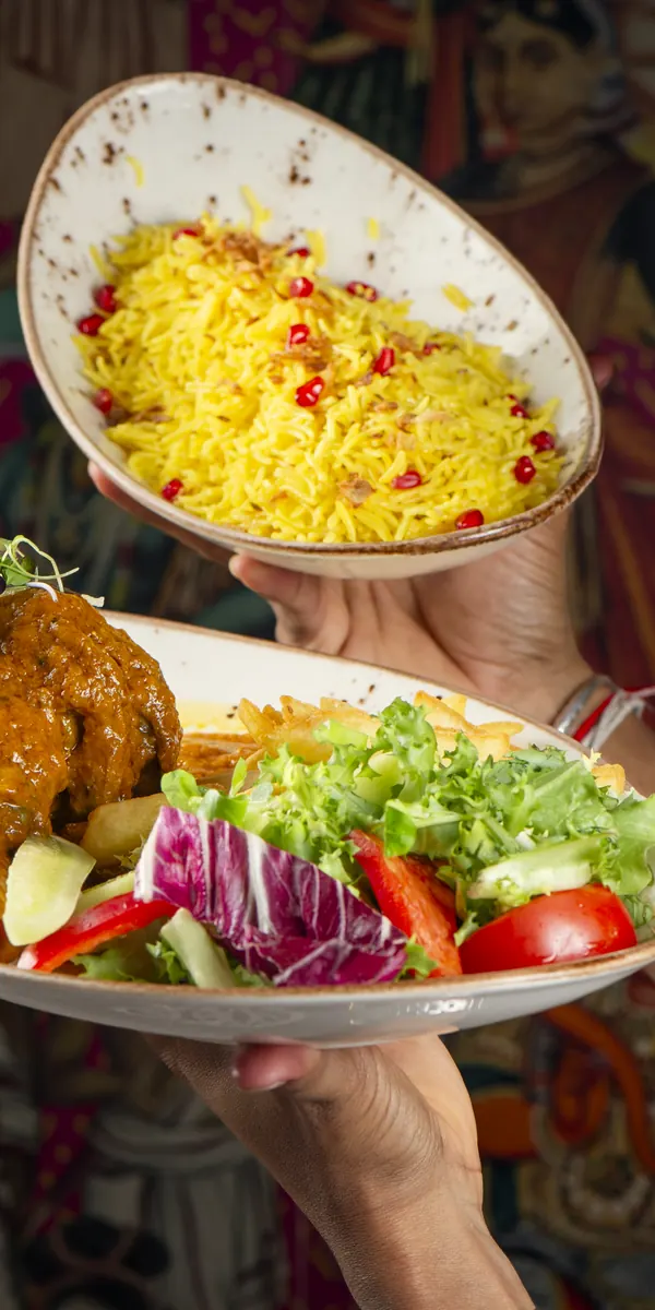 A bowl of yellow rice with pomegranate held up and, below it, a plate of lamb with salad, in front of an Indian mural