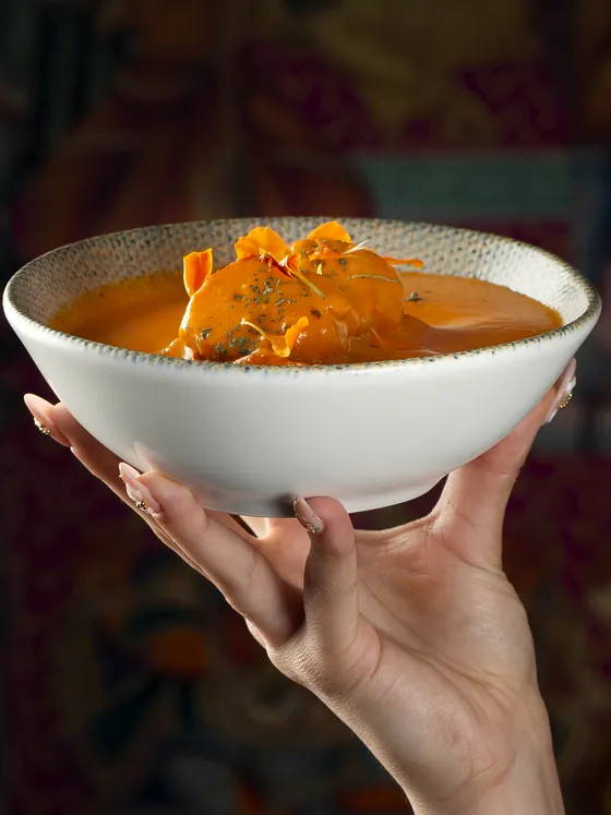 Chicken tikka masala with marigold petals, in a bowl held up in front of an Indian mural